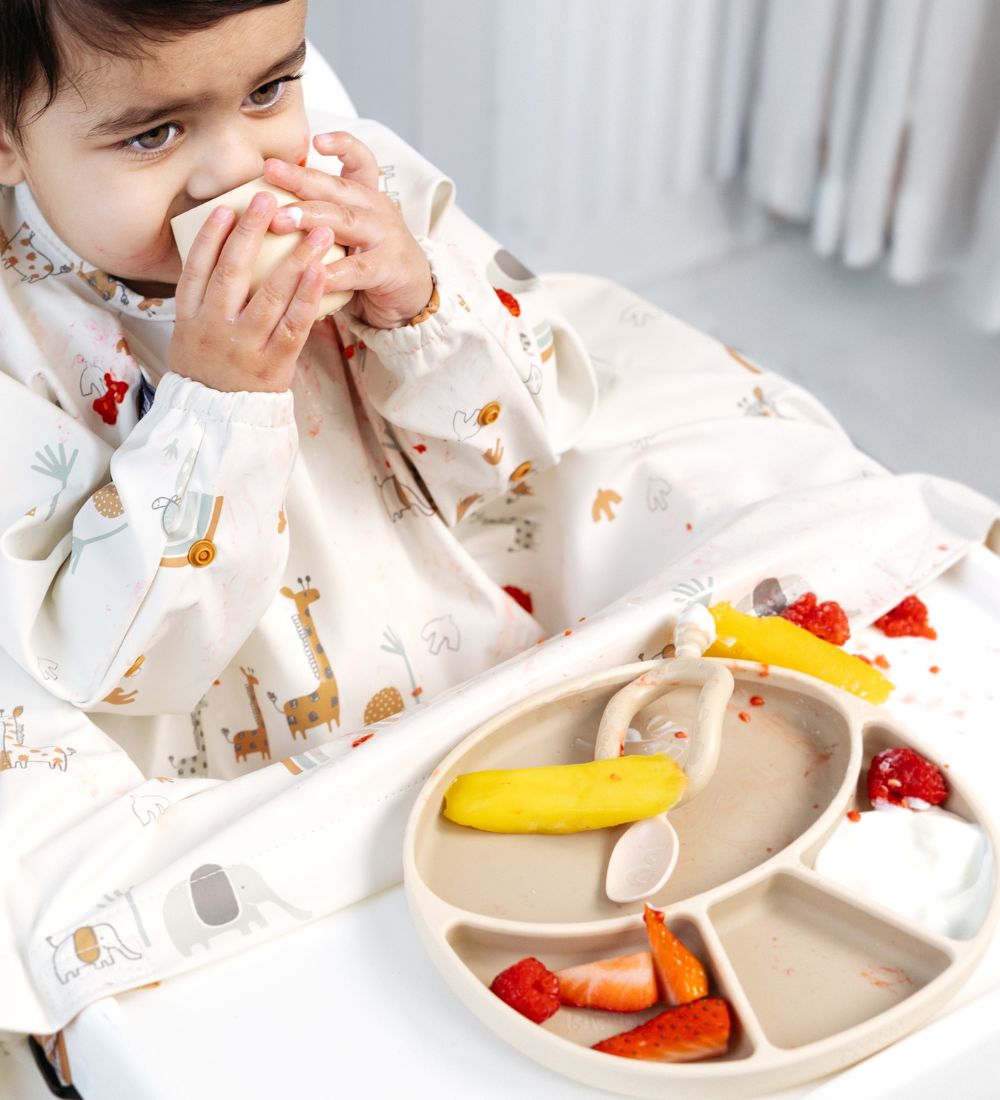 Baby in a long-sleeved animal print bib, holding a cup, with a divided plate of fruit and yoghurt.