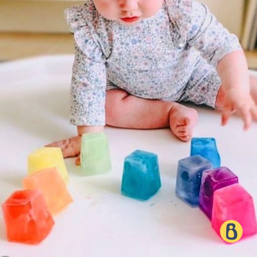 A baby playing with colourful rainbow ice cubes on a white surface.