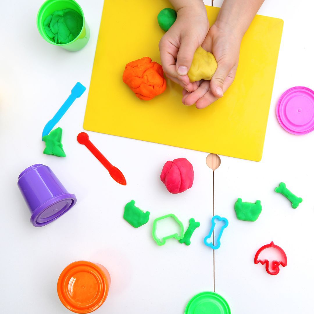 Child's hands playing with yellow gluten-free playdough, surrounded by colourful cutters and pots.