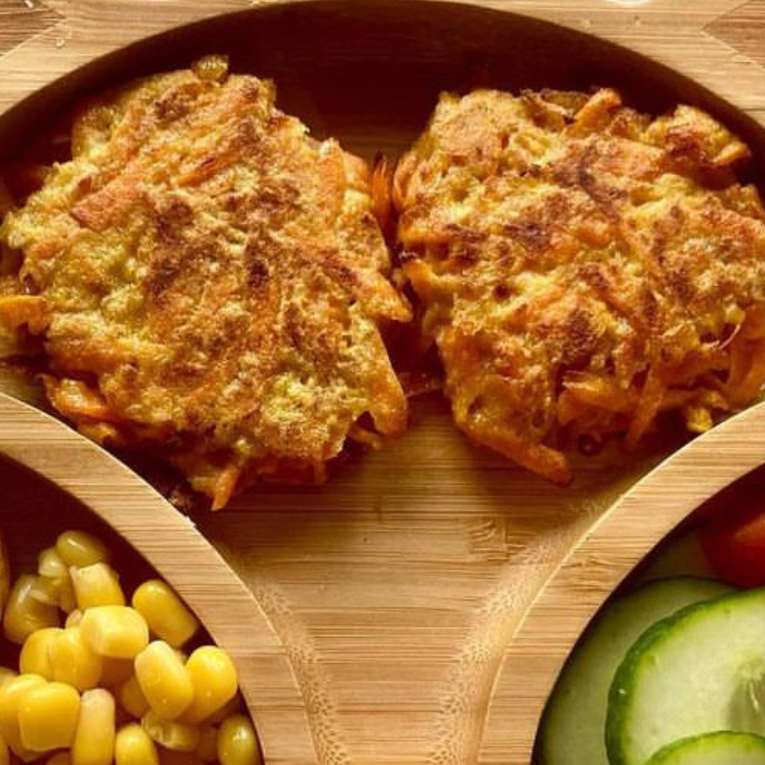 Two golden-brown carrot fritters on a wooden plate with sweetcorn and cucumber slices.