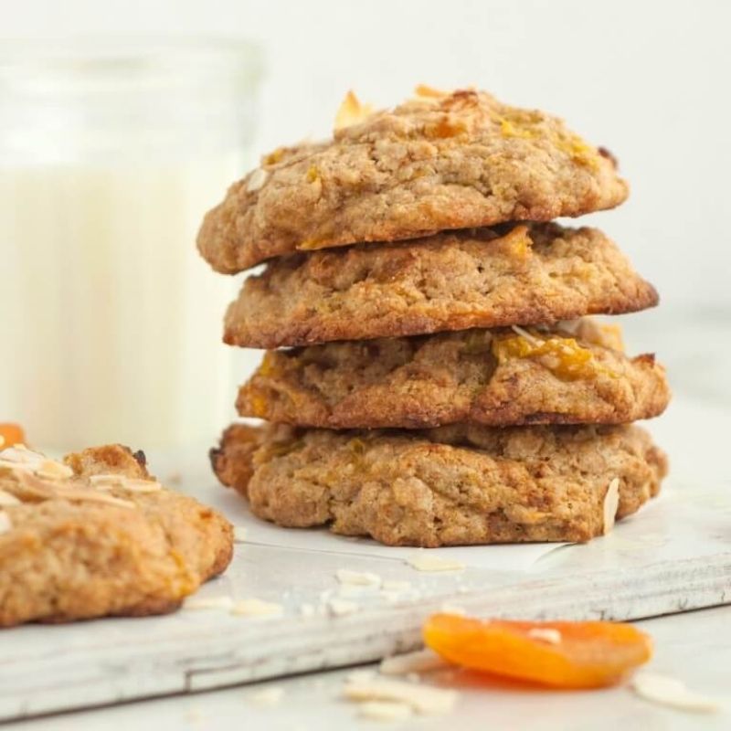 Stack of four golden-brown apricot cookies with flaked almonds, next to a glass of milk.
