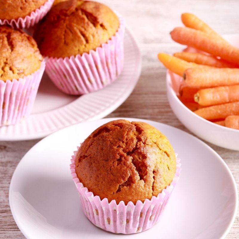 Golden carrot muffins in pink cases on white plates, with fresh carrots in a bowl.