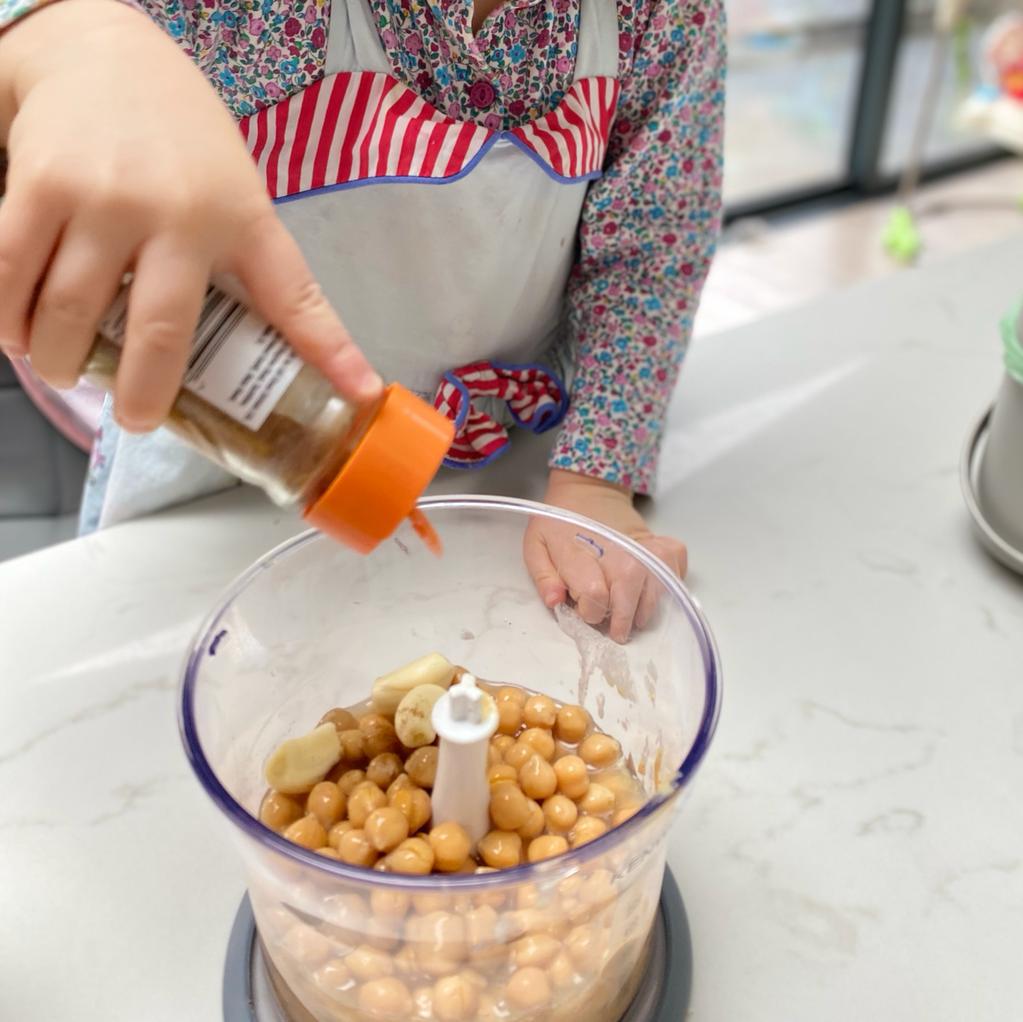 Child in floral top and apron adding spices to chickpeas and garlic in a food processor.