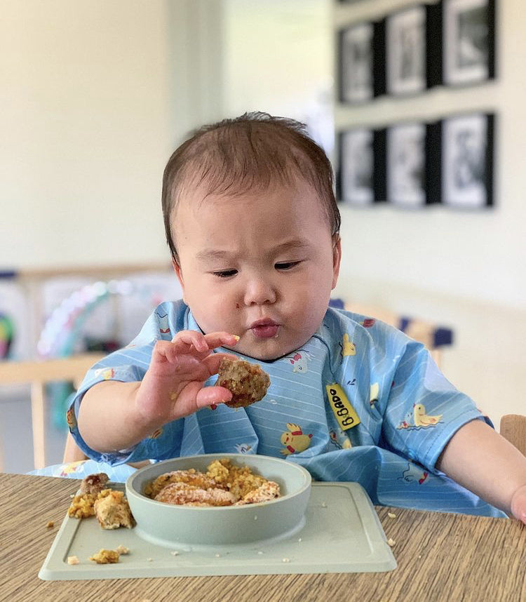Baby in a blue bib eating a meatball from a bowl on a highchair tray.