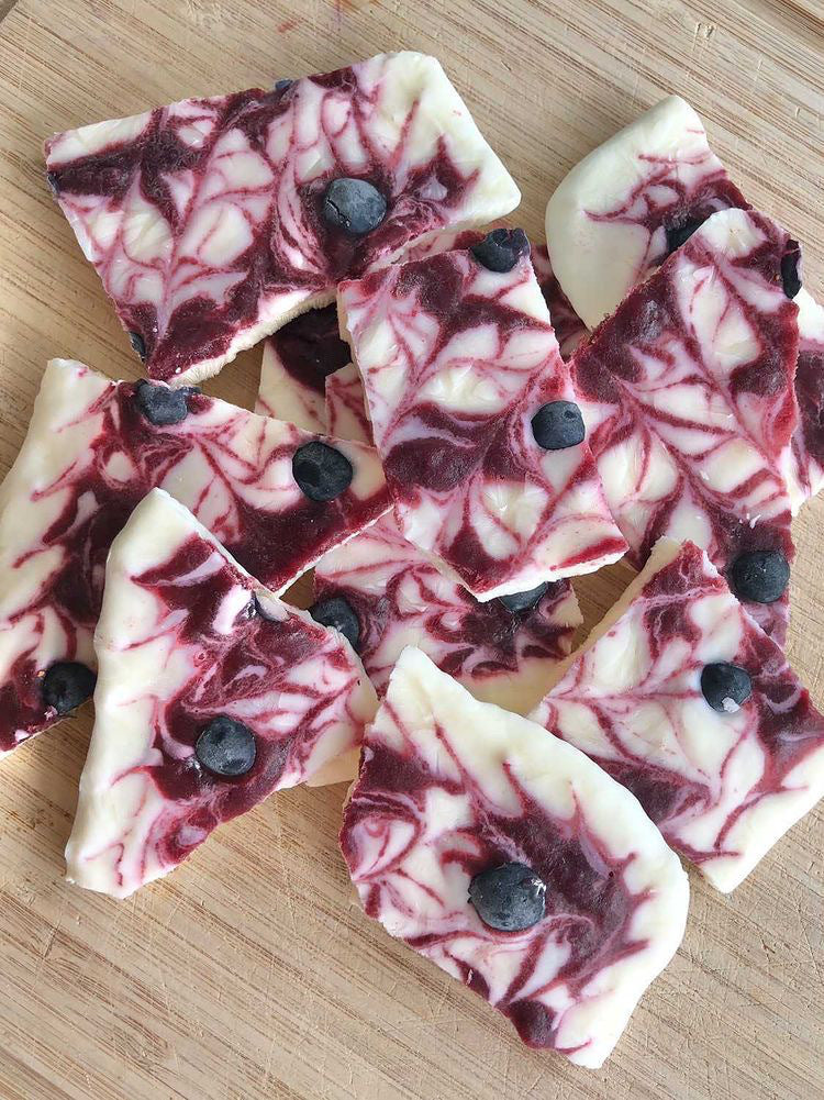 White frozen yoghurt bark with purple blueberry swirls and whole blueberries on a wooden board.
