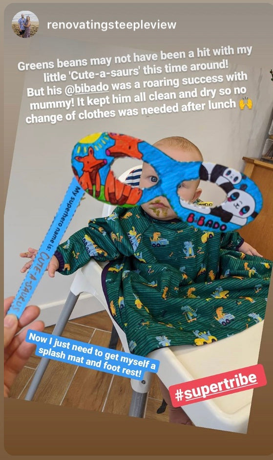Baby in a green dinosaur bib, holding a colourful 'Cute-a-saurus' superhero mask.