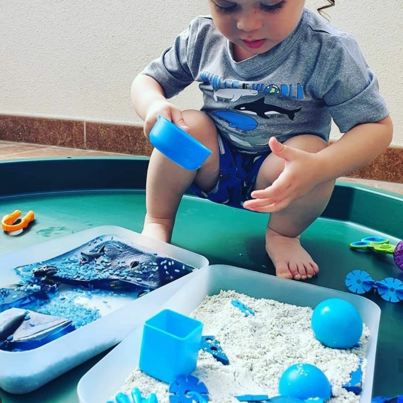 Toddler playing with blue moonsand and accessories in a green sensory bin.