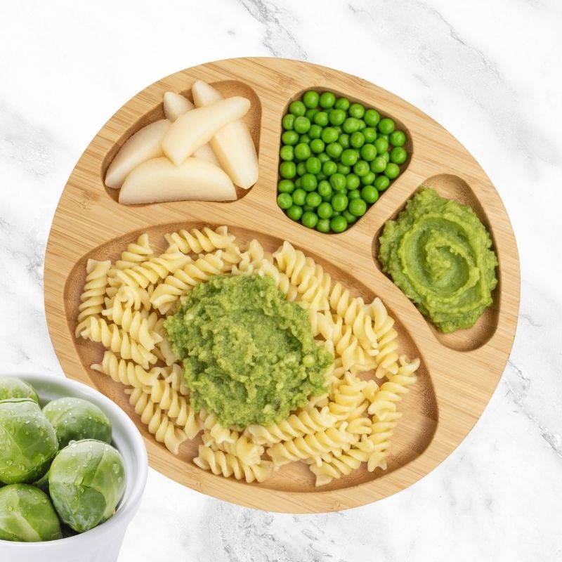 Baby-led weaning meal: pear slices, peas, and pasta with pea and brussel sprout purée on a bamboo plate.
