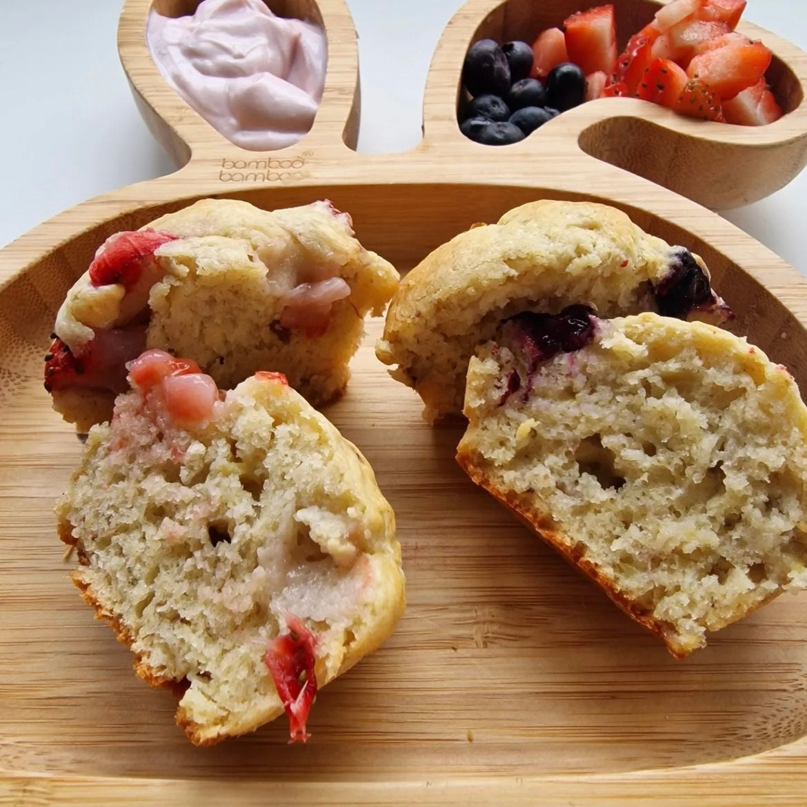Two banana muffins, one with strawberries, cut in half on a wooden bunny plate with fruit and yoghurt.