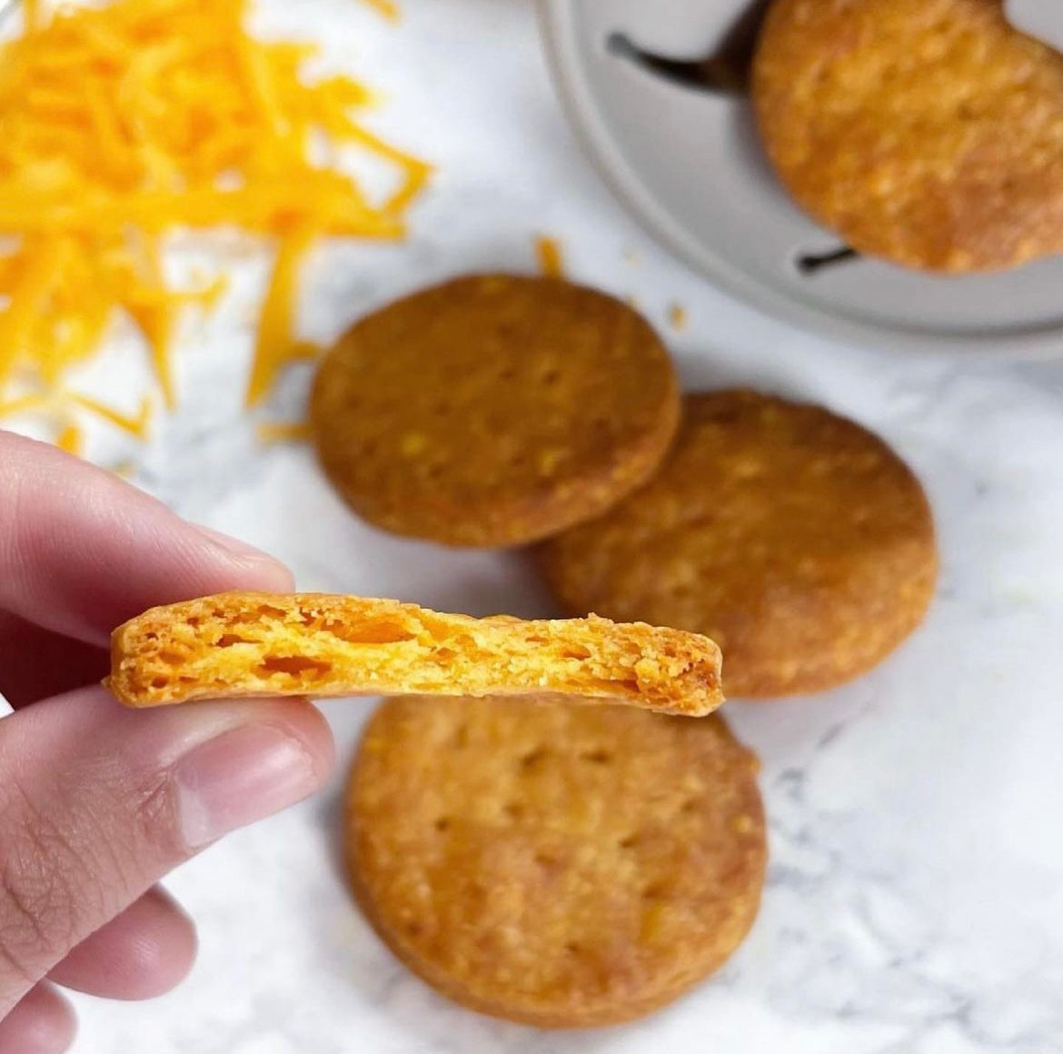 Hand holding a golden, flaky homemade cheese biscuit, with more biscuits and grated cheddar in the background.