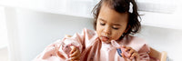 Toddler in a pink Bibado bib, eating with a matching spoon.