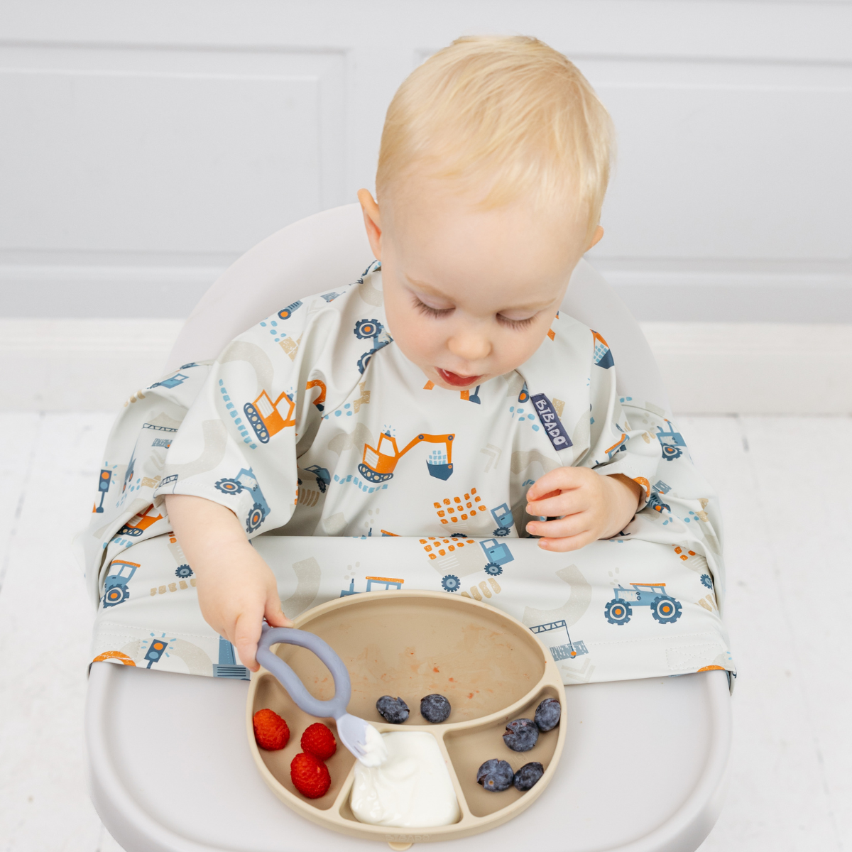 Blonde toddler in a short-sleeve, light gray coverall bib with orange construction vehicle print, eating from a beige divided plate. - Product image