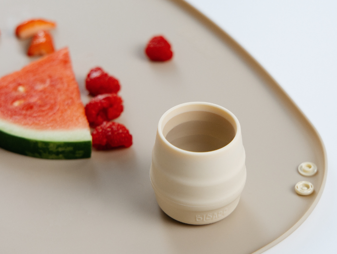A beige silicone cup, shown on a matching beige mat with a watermelon slice and raspberries. - image