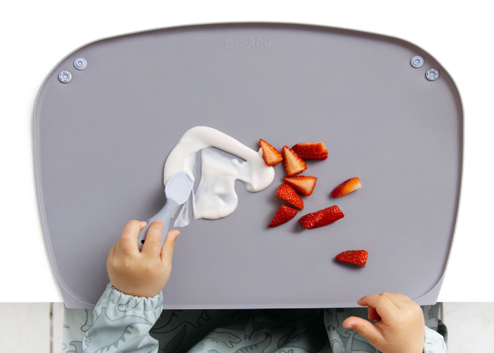 A child wearing a light blue dinosaur-print bib sits in a high chair, using a blue-greyu silicone feeding mat to scoop a smear of white yogurt with a blue spoon. Sliced strawberries are scattered on the tray. - product info image