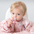 Blonde baby in a pink floral bib, holding a pink silicone Dippit™ spoon with food on it.
