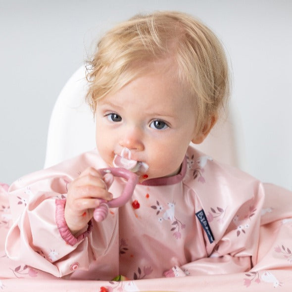 Blonde baby in a pink floral bib, holding a pink silicone Dippit™ spoon with food on it. - Product image