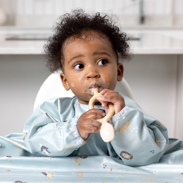Baby in a light blue bib, holding a beige Dippit™ dual-ended spoon and dipper. - Product image