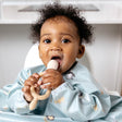 Baby in a blue patterned bib holding a beige Dippit™ multi-stage baby spoon and dipper.