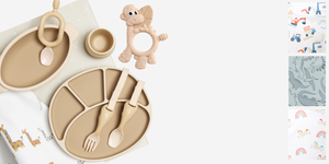 Neutral-toned baby feeding set with silicone plates, bowl, cutlery, and a dinosaur teether, beside patterned fabrics.