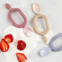 Three silicone Dippit™ or Sporkit™ baby spoons in pink, beige, and lavender, surrounded by yogurt and strawberries.