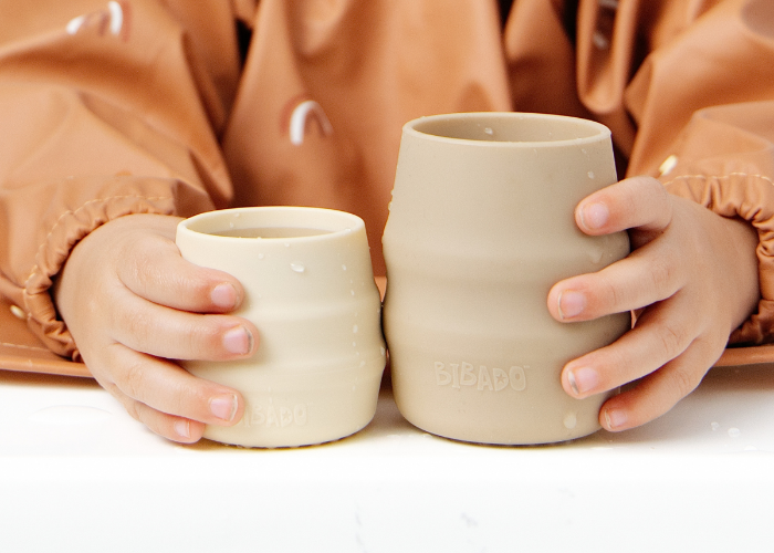 Two beige silicone cups, one smaller than the other, held by a child wearing a brown Coverall bib. The child's hands and torso are visible, and the cups are shown against a clean white background. - product info image