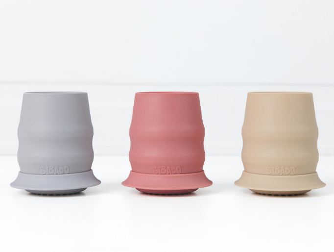 Three silicone baby cups with stabilising skirts in blue-grey, dusty pink, and beige, arranged side by side on a clean white surface. - Product image