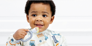 Smiling baby in a patterned bib holding a grey teether with yellow food.