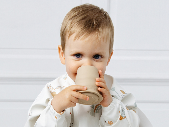 A baby wearing a white long-sleeve bib with a safari animal print is drinking from a beige silicone cup. - image