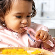 Baby in a pink floral bib uses a pink Dippit™ multi-stage spoon to dip food from a bamboo plate.