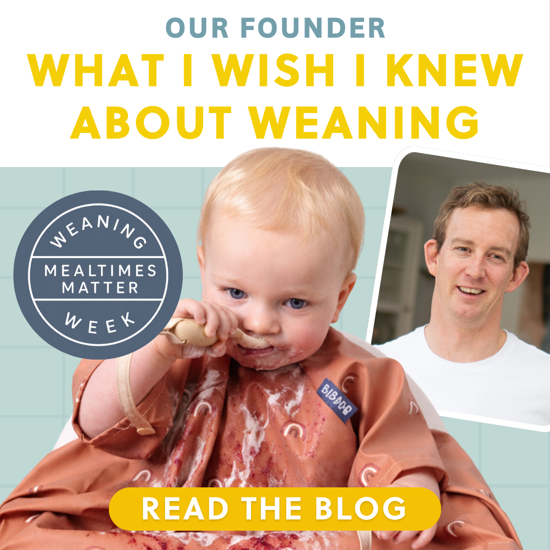 Blonde baby in a messy terracotta bib eating with a wooden spoon, next to a smiling man.
