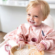 Happy baby in a pink floral bib, covered in yogurt, holding a wooden bowl and pink Dippit™ spoon.