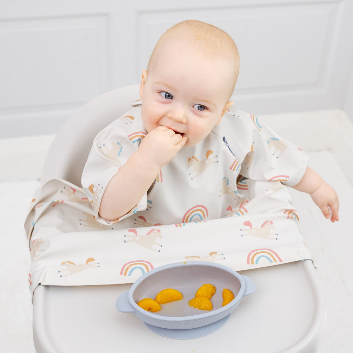 Baby in a white, short-sleeve coverall bib with unicorn and rainbow print, eating from a gray bowl. - Product image