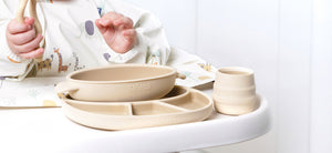 Baby in a safari-print bib with a beige silicone bowl, divided plate, and cup on a highchair.