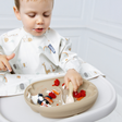 A baby wearing a white bib with a safari animal print sits in a high chair, reaching for food on a beige silicone plate.