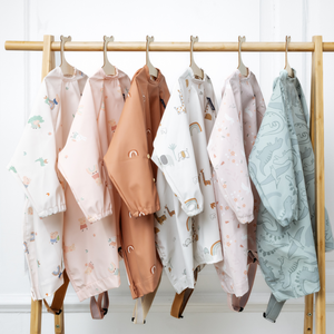A row of long-sleeved baby bibs in various pastel colours and animal prints hanging on a wooden rail.
