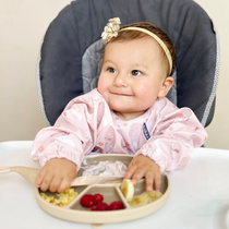 Smiling baby in a pink long-sleeved bib, sitting in a high chair with a plate of food.