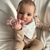 Baby in beige outfit and patterned bib chewing a pink dinosaur teether.