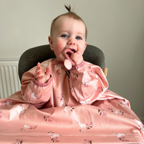 Happy baby in a high chair, wearing a pink long-sleeved bib with a white unicorn pattern, holding a spoon.