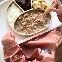 Toddler in a peach bib eating porridge, banana, raisins, and yoghurt from a beige silicone plate.