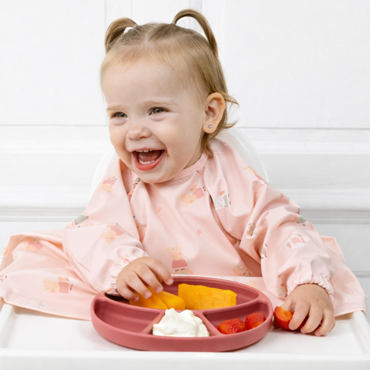 Child in a pink Coverall bib smiling whilst eating fruit from a blush silicone plate. - Product image