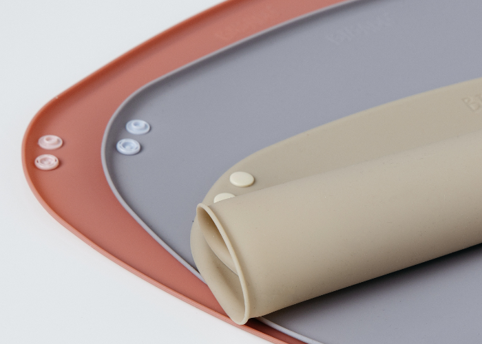 Close-up of a beige rolled silicone weaning mat with snap buttons resting on a blue-grey version fully rolled out, which is layered again over a pink mat on a clean white background. - product info image
