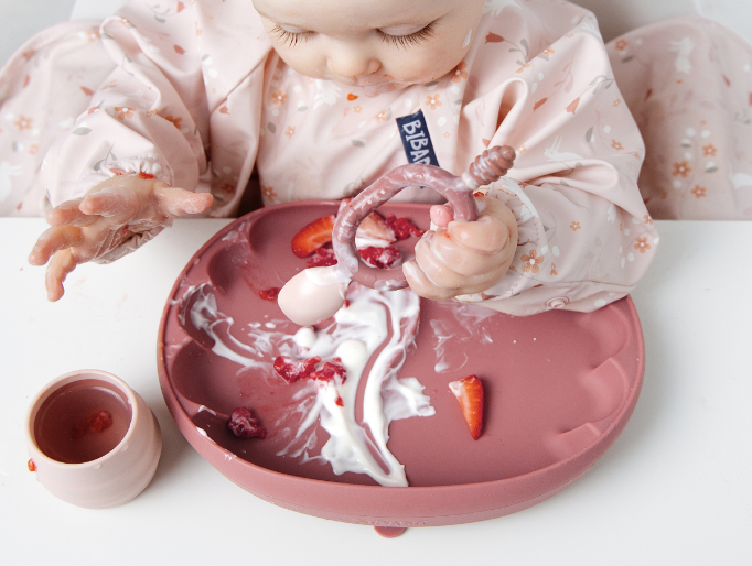 A baby wearing a light pink patterned bib sits in a high chair and eats messy food from a dusty pink silicone plate. The baby holds a pink Dippit spoon. - image