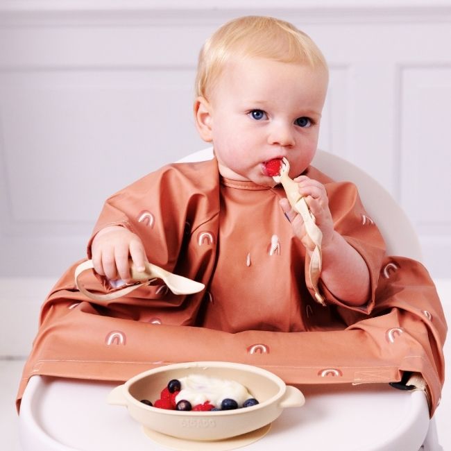 Blonde baby in a terracotta waterproof bib with rainbow print, eating berries and yoghurt from a beige bowl.