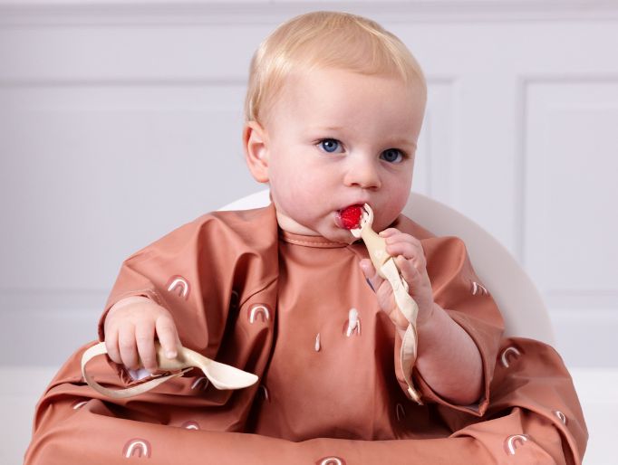Blonde baby in a terracotta rainbow print coverall bib, holding wooden spoons and eating a strawberry. - image