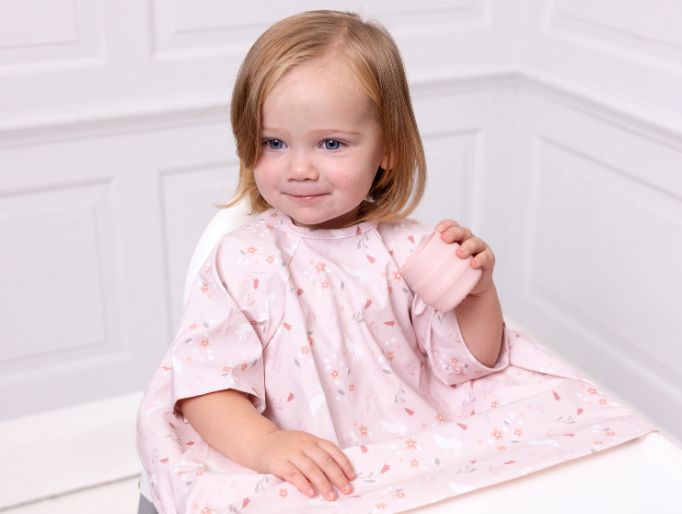 Toddler in a pink floral coverall bib with long sleeves, holding a matching pink cup. - image