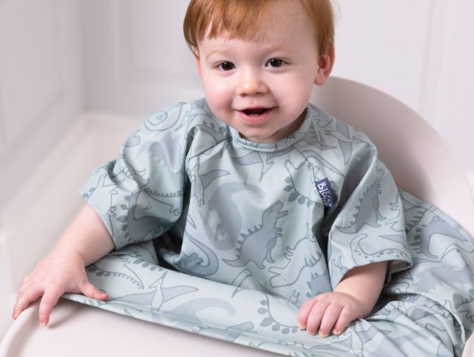 Smiling baby in a light blue dinosaur-print coverall bib with short sleeves, sitting in a highchair. - image