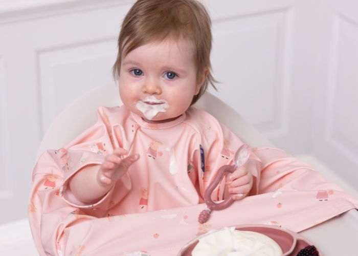 Baby in a pink, long-sleeved coverall bib with food on her face, holding a spoon. - product info image