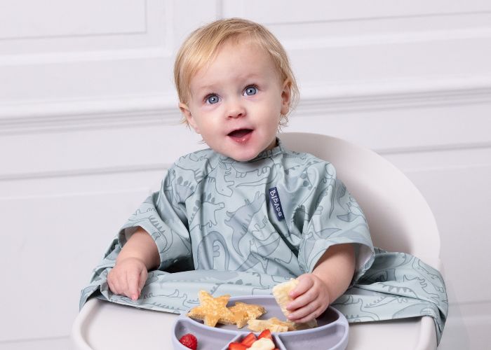 Happy toddler in a grey dinosaur print coverall bib, eating snacks from a purple suction plate. - product info image