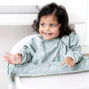 Smiling baby in a long-sleeved, light green dinosaur-patterned coverall bib in a highchair.
