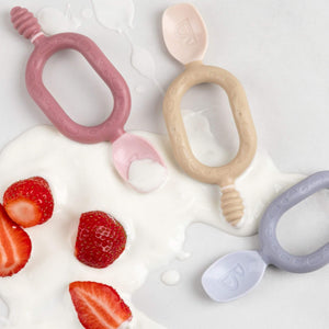 Three colourful silicone baby spoons with textured handles in spilt yoghurt and strawberries.